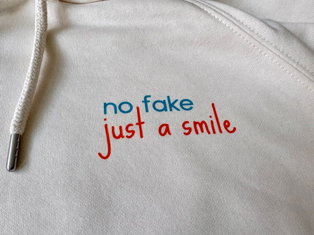 Detailaufnahme des nachhaltigen Bio-Hoodies von der Kieler Künstlerin Diana Gumz. Auf dem cremeweißen Stoff ist der aufgedruckte, farbige Schriftzug "no fake just a smile" neben der Kapuzenkordel zu sehen.