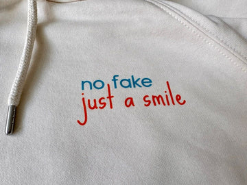 Detailaufnahme des nachhaltigen Bio-Hoodies von der Kieler Künstlerin Diana Gumz. Auf dem cremeweißen Stoff ist der aufgedruckte, farbige Schriftzug "no fake just a smile" neben der Kapuzenkordel zu sehen.
