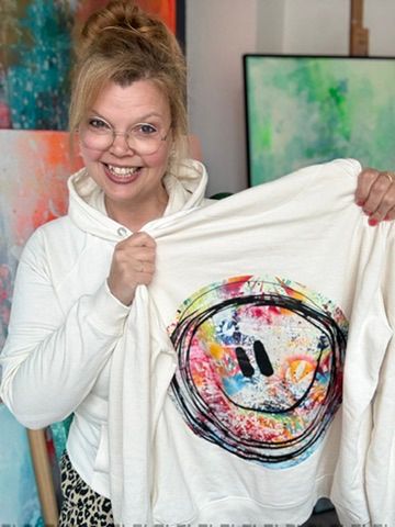 Die Kieler Künstlerin Diana Gumz lächelt in ihrem Atelier, während sie einen cremeweißen, nachhaltigen Bio-Hoodie mit dem handillustrierten, farbenfrohen Smiley-Kunstwerk-Print auf der Brust präsentiert.