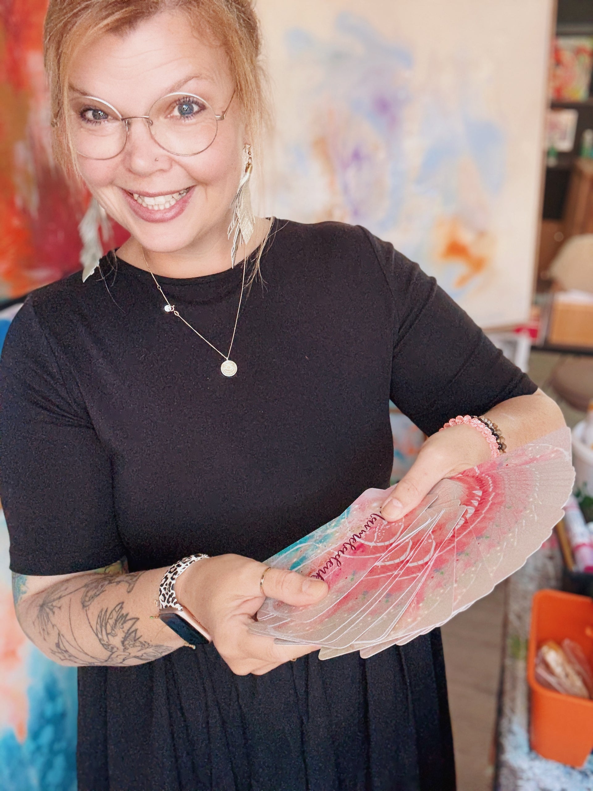 Die lächelnde Kieler Künstlerin Diana Gumz hält ihr handillustriertes Farbkartenset für Achtsamkeit und Affirmationen in den Händen. Im Hintergrund sind ihre abstrakten Gemälde im Atelier zu sehen.