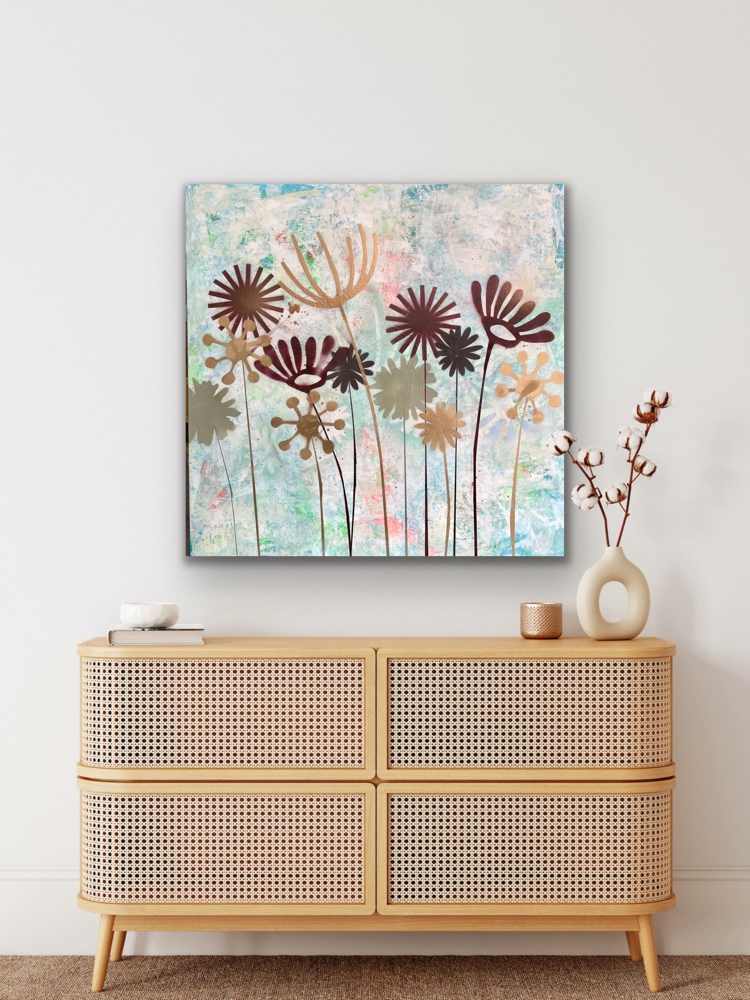 Einrichtungsbeispiel im natürlichen Boho-Stil: Das abstrakte Blumengemälde 'Summertime' hängt rahmenlos über einer hellen Rattan-Kommode. Daneben steht eine skulpturale Vase mit Baumwollzweigen.