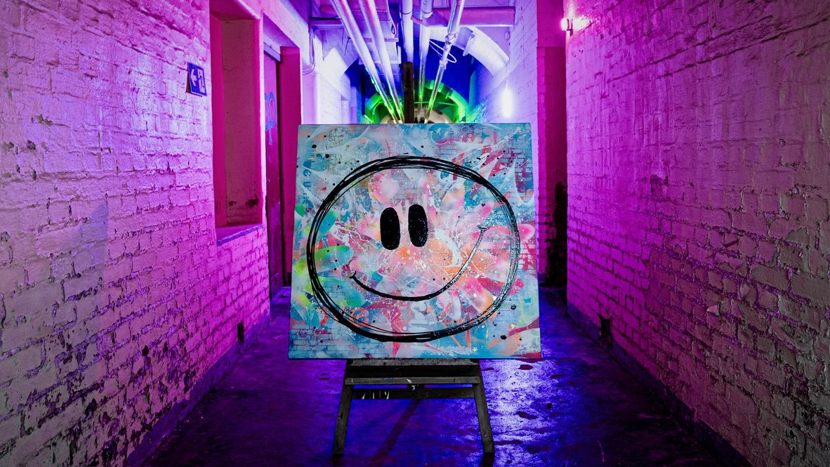 Inszenierung wie in einer Galerie: Das bunte Gemälde 'Big Smile' steht auf einer dunklen Holzstaffelei mitten in einem langen, alten Backsteingang, der komplett mit pinkem und lila Neonlicht ausgeleuchtet ist