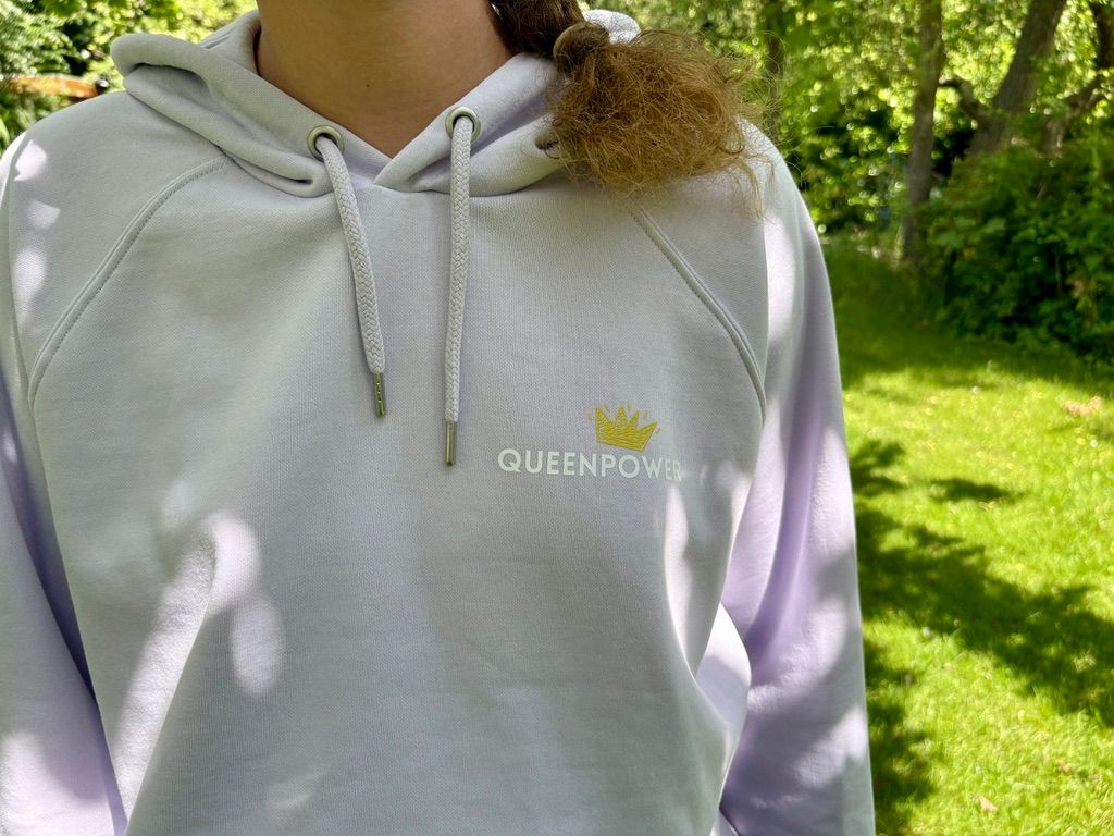 Nahaufnahme der Vorderseite des fliederfarbenen Hoodies im Freien. Auf der linken Brust leuchtet ein detailreicher Print: Eine kleine goldene Krone mit dem Schriftzug 'QUEENPOWER'.