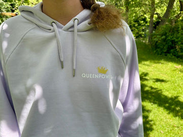 Nahaufnahme der Vorderseite des fliederfarbenen Hoodies im Freien. Auf der linken Brust leuchtet ein detailreicher Print: Eine kleine goldene Krone mit dem Schriftzug 'QUEENPOWER'.