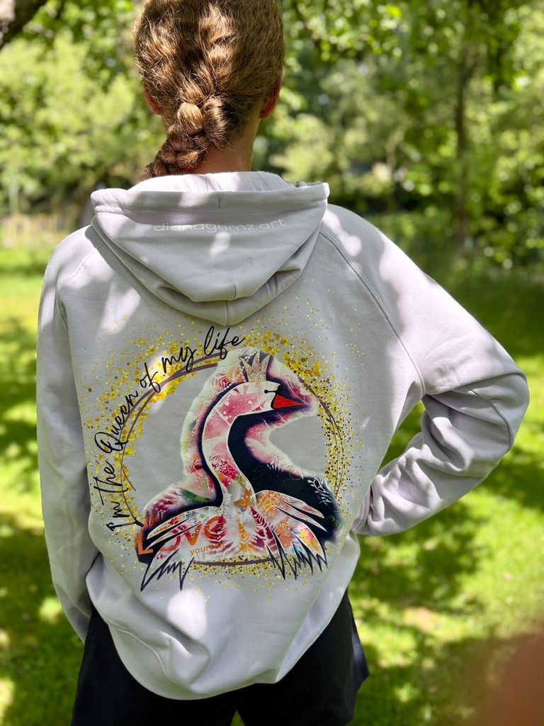 Eine Person steht von hinten fotografiert in einem sonnigen Garten und trägt den fliederfarbenen Hoodie 'Schwanenkönigin'. Auf dem Rücken leuchtet der große, bunte Kunst-Print eines Schwans mit Krone, umrahmt von dem goldenen Schriftzug 'I'm the Queen of my life' und 'LOVE yourself'.