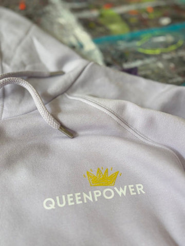 Nahaufnahme des fliederfarbenen Hoodies 'Schwanenkönigin'. Der Fokus liegt auf dem kuscheligen Stoff und dem Brust-Print: Eine gelb-goldene Krone mit dem weißen Schriftzug 'QUEENPOWER'. Im unscharfen Hintergrund ist ein buntes abstraktes Gemälde zu erahnen.