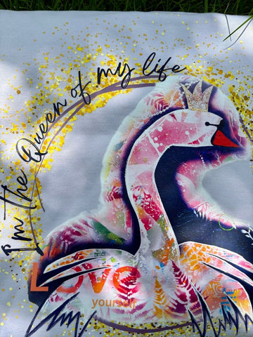 Detailaufnahme des großen, bunten Schwanen-Kunstprints auf dem Rücken des fliederfarbenen Hoodies im Sonnenlicht. Das Motiv zeigt einen Schwan mit Krone und die Schriftzüge 'I'm the Queen of my life' und 'LOVE yourself', umgeben von funkelnden goldenen Glitzer-Details.