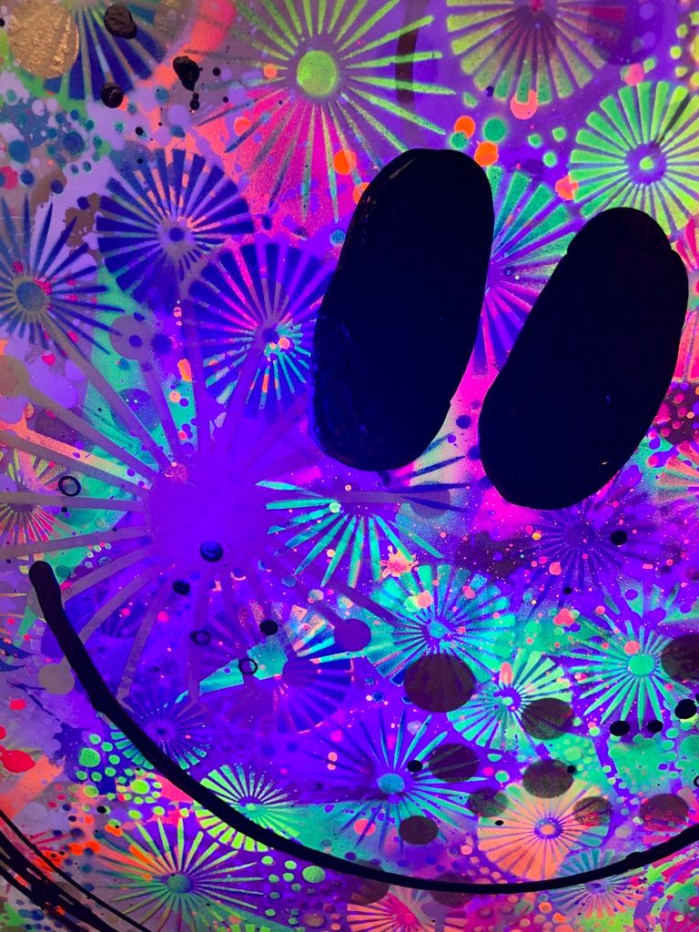 Nahaufnahme des Gemäldes 'Summer Smile' unter Schwarzlicht (UV-Licht). Die kreisförmigen Muster leuchten intensiv in fluoreszierendem Neon-Grün, Neon-Pink und Orange auf einem dunklen violett-blauen Hintergrund. Die schwarzen Augen des Smileys heben sich dunkel ab.