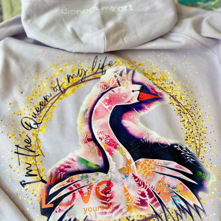 Nahaufnahme der Rückseite des fliederfarbenen Hoodies. Zu sehen ist das farbenfrohe Pop-Art Schwanen-Motiv mit der goldenen Aufschrift 'I'm the Queen of my life'. Im oberen Bereich ist die Kapuze des Pullovers zu erkennen.
