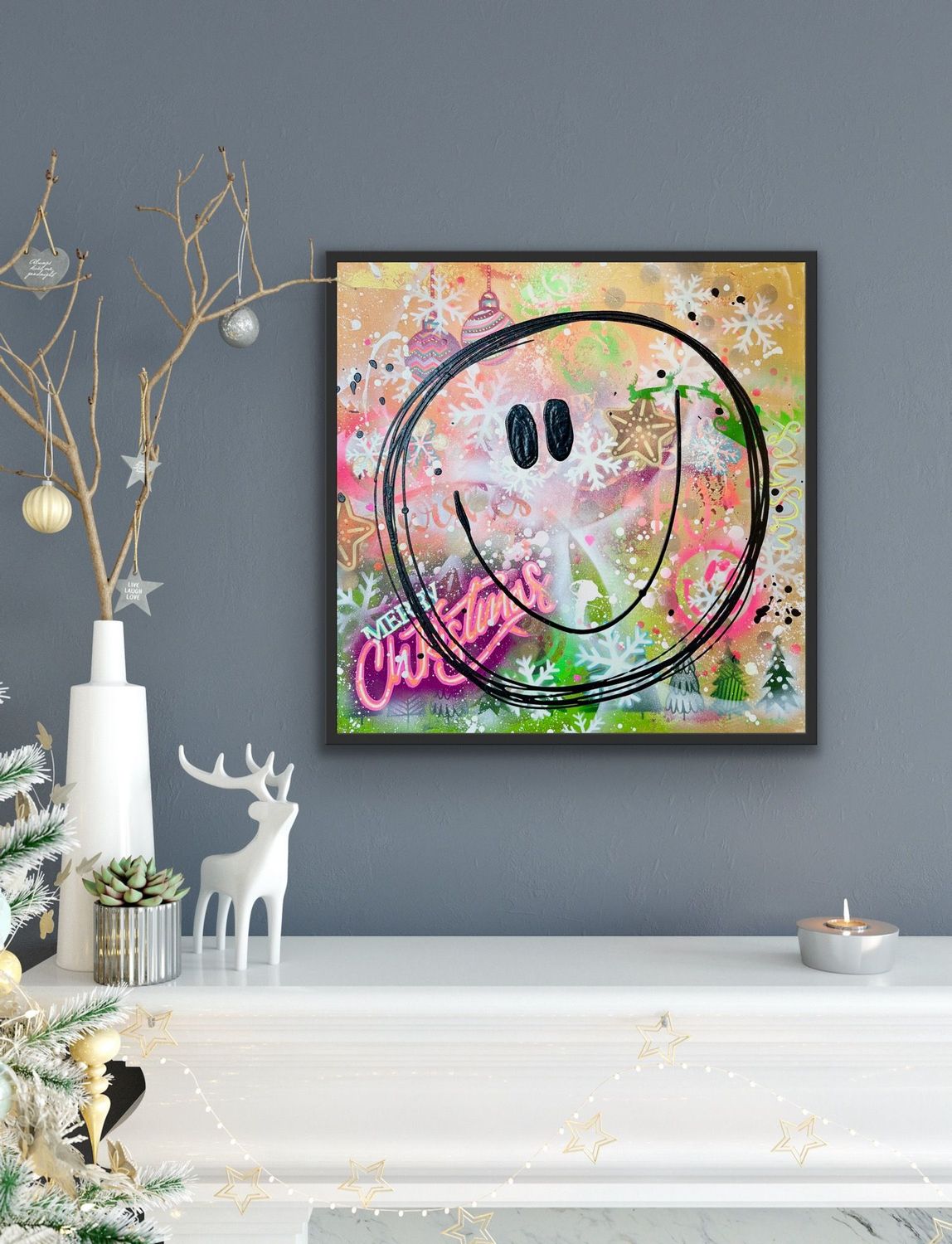Einrichtungsbeispiel für moderne Weihnachtsdeko: Das bunte Gemälde 'Christmas Smile' hängt in einem schwarzen Rahmen an einer grauen Wand über einer weißen Kamin-Konsole, dekoriert mit einem goldenen Teelicht und einer weißen Rentier-Figur.